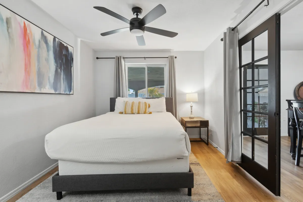 A tranquil bedroom with white bedding and a yellow accent pillow, abstract wall art, a modern ceiling fan, lampside table, and curtains framing a window. The space feels airy, quiet, and designed for restful moments.