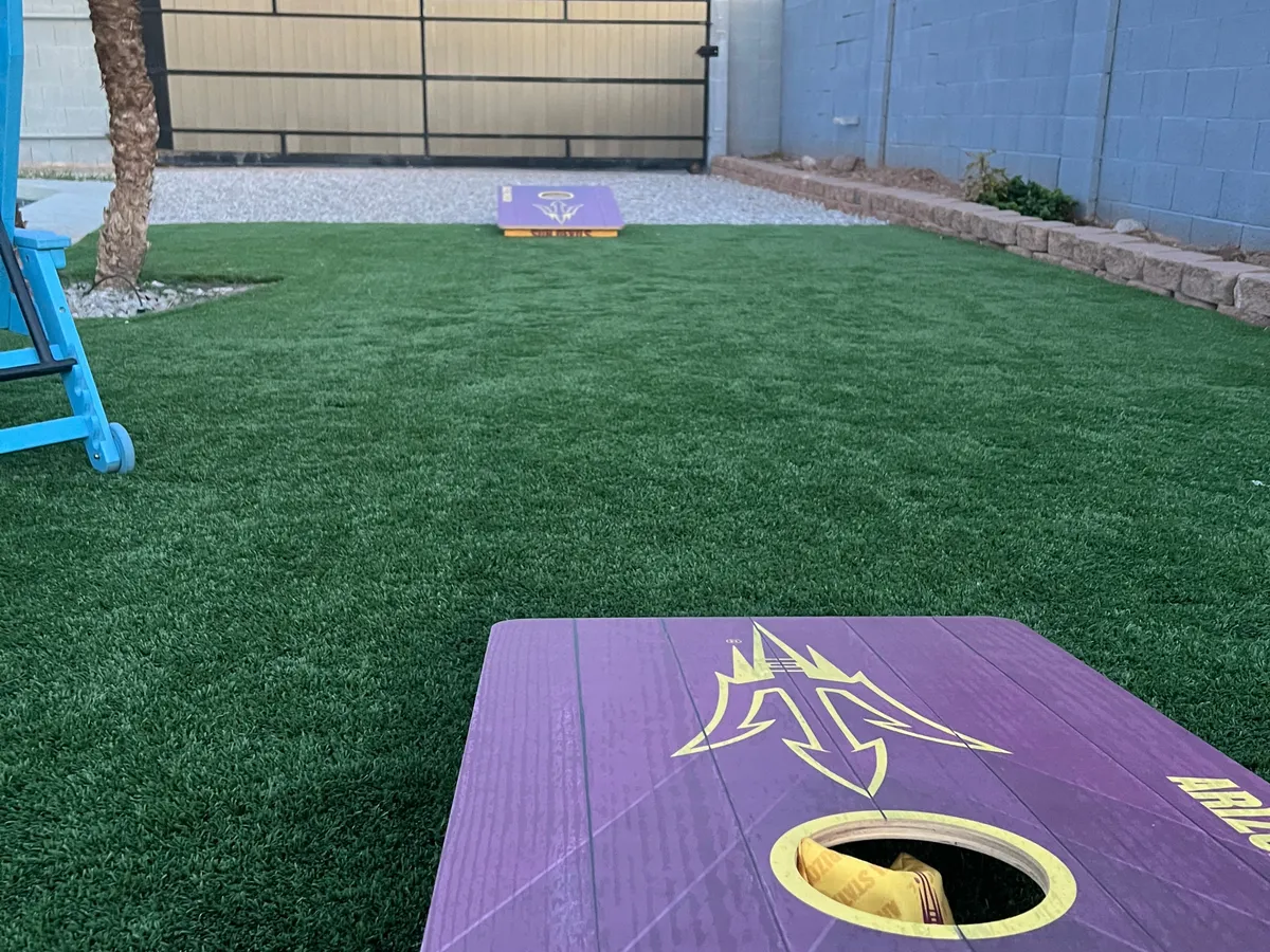A playful backyard space with cornhole boards set on artificial grass, shaded by a palm tree, and complemented by a gravel pathway. The area is designed for games and casual socializing.