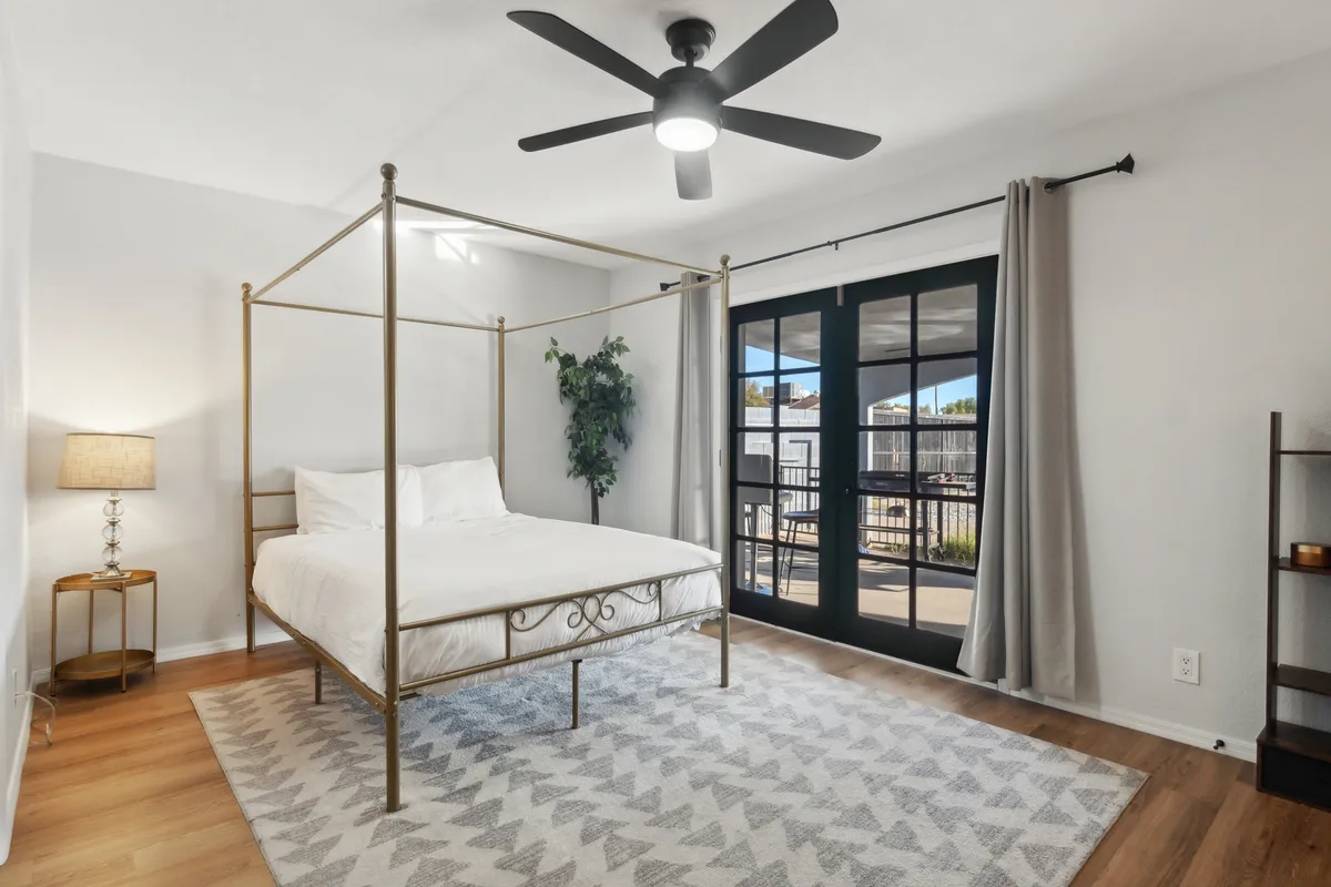 A serene bedroom featuring a canopy bed with plush bedding, large windows letting in natural light, a decorative lamp, green plant, and area rug. The atmosphere is calm and inviting, ideal for relaxation and restful sleep.