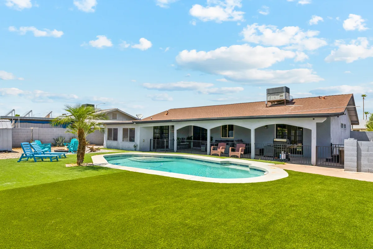 The hero image features a sparkling swimming pool surrounded by lush green lawn and swaying palm trees, with comfortable outdoor seating arranged for relaxing and entertaining. The scene is bathed in warm Arizona sunlight, suggesting opportunities for swimming, sunbathing, and enjoying meals or drinks by the pool in a private and inviting setting.