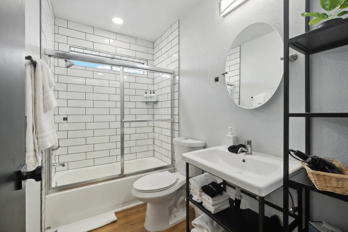 A fresh and inviting bathroom with a glass shower enclosure, vanity with sink, round mirror, and shelves stocked with towels. The space is bright, modern, and well-appointed for relaxation or post-pool convenience.