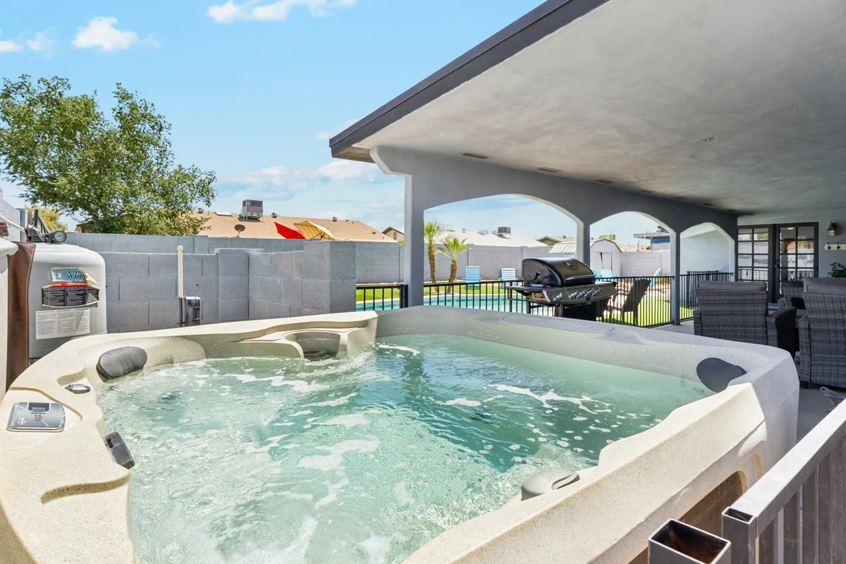 This exterior image features a well-appointed patio with a hot tub, grill, and comfortable seating. The area is surrounded by lush greenery and is designed for relaxation and socializing, offering a refreshing outdoor escape.