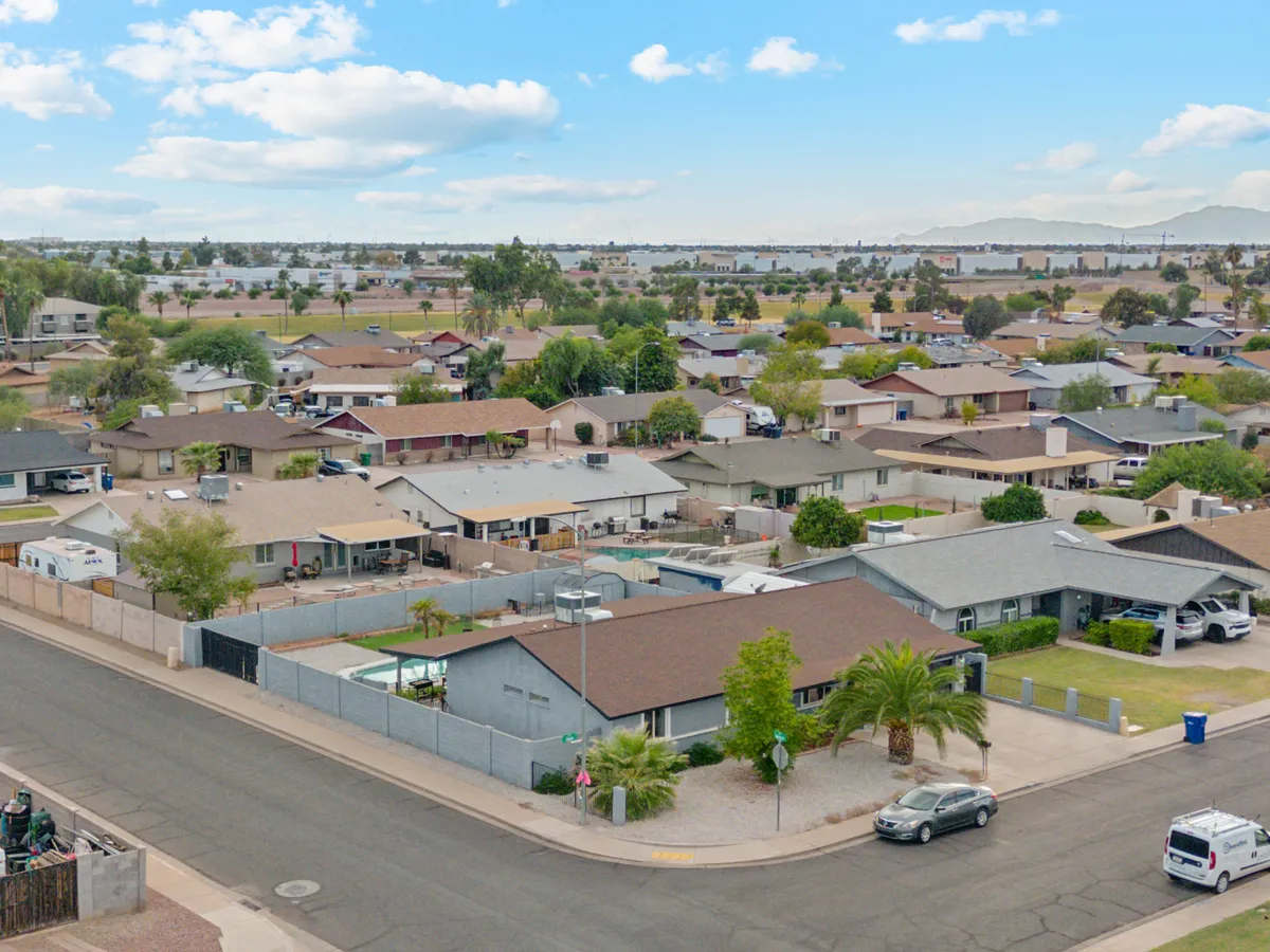 A broad exterior view shows a residential street lined with green and palm trees, featuring a shared swimming pool in the foreground. The scene feels active and family-friendly, evoking neighborhood gatherings, children playing, and inviting outdoor moments.